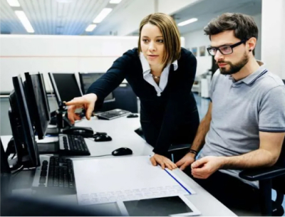 White female IT manager pointing something out on a computer screen to white male IT worker